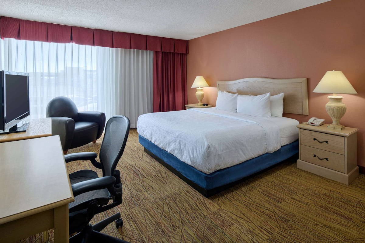 A hotel room with a bed, two bedside lamps, a desk with a chair, a TV, and a window with curtains.