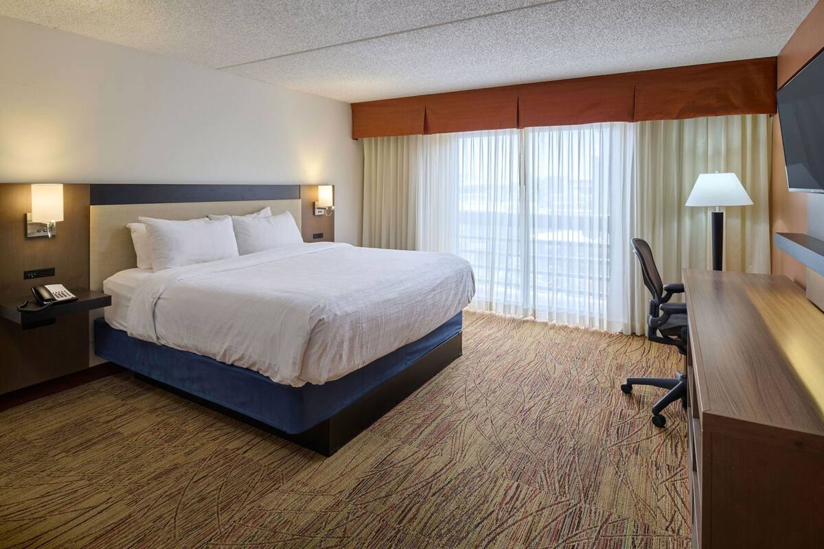 A hotel room with a large bed, a desk with a chair, a TV, and a window with curtains.