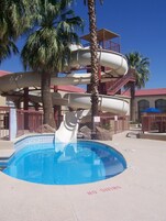 Estrutura de toboáguas com piscina na base, palmeiras e um edifício com telhado vermelho ao fundo.