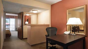 Habitación de hotel con escritorio, silla y un espejo grande. Se observa una cama al fondo y una puerta que da a otra habitación.