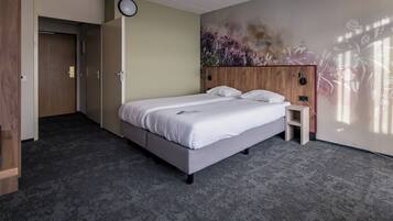 A hotel room with a bed, a nightstand, a wall-mounted lamp, and a door leading to another room.