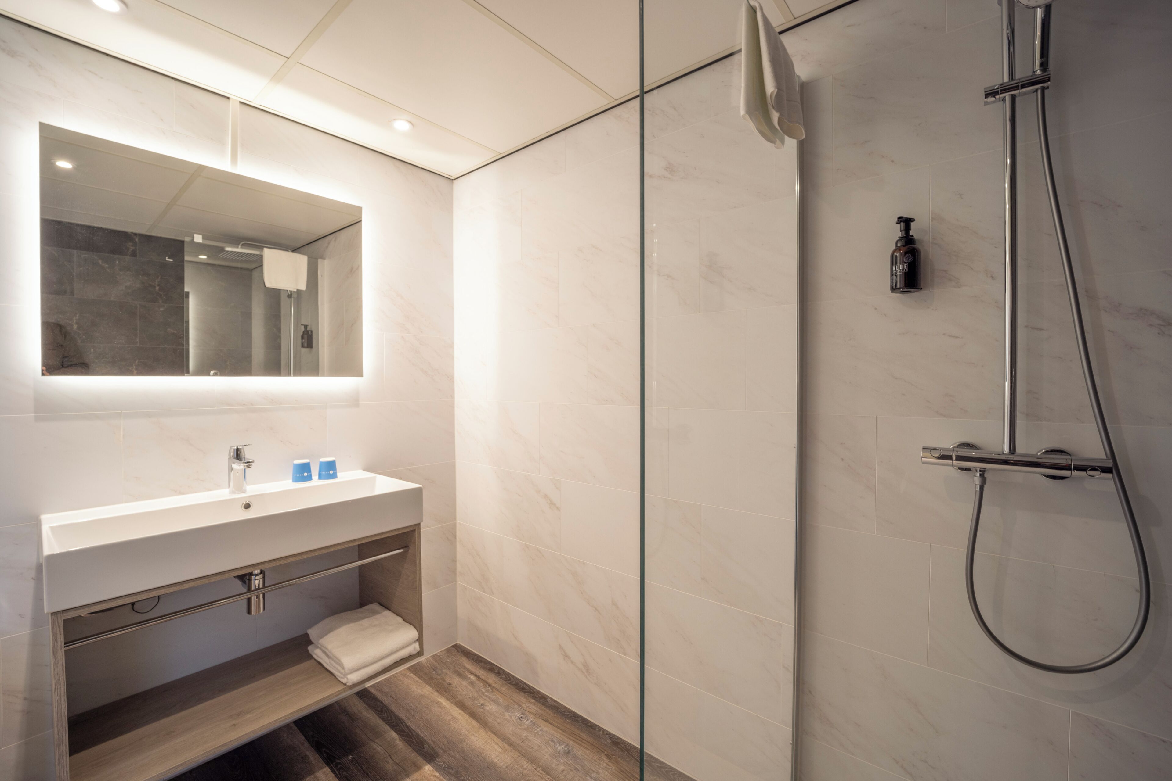 A modern bathroom with a glass shower enclosure, a white vanity with a mirror, and a wooden floor.
