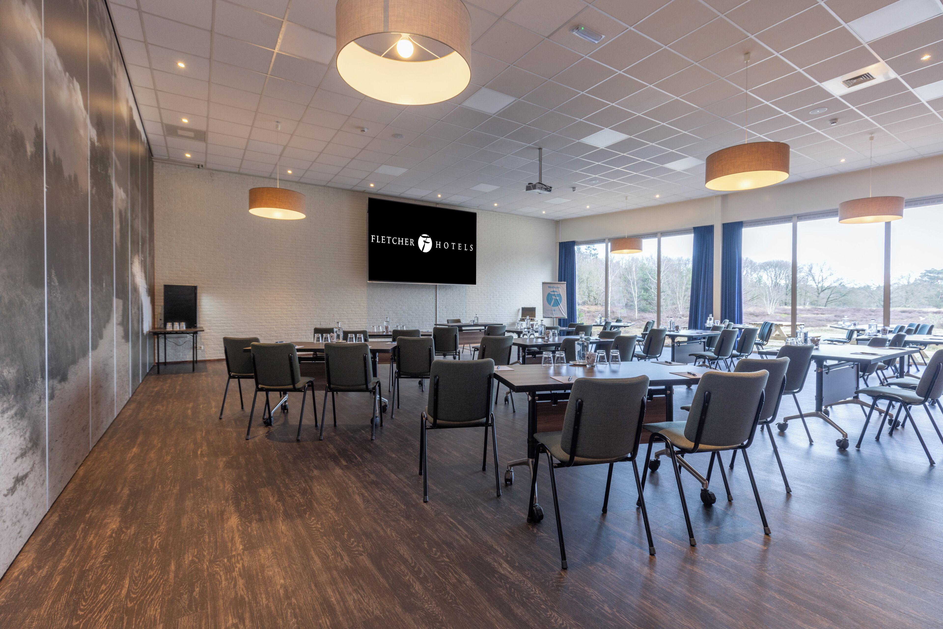 A conference room with tables and chairs, a large screen displaying 'Fletcher Hotels', and a view of trees through large windows.