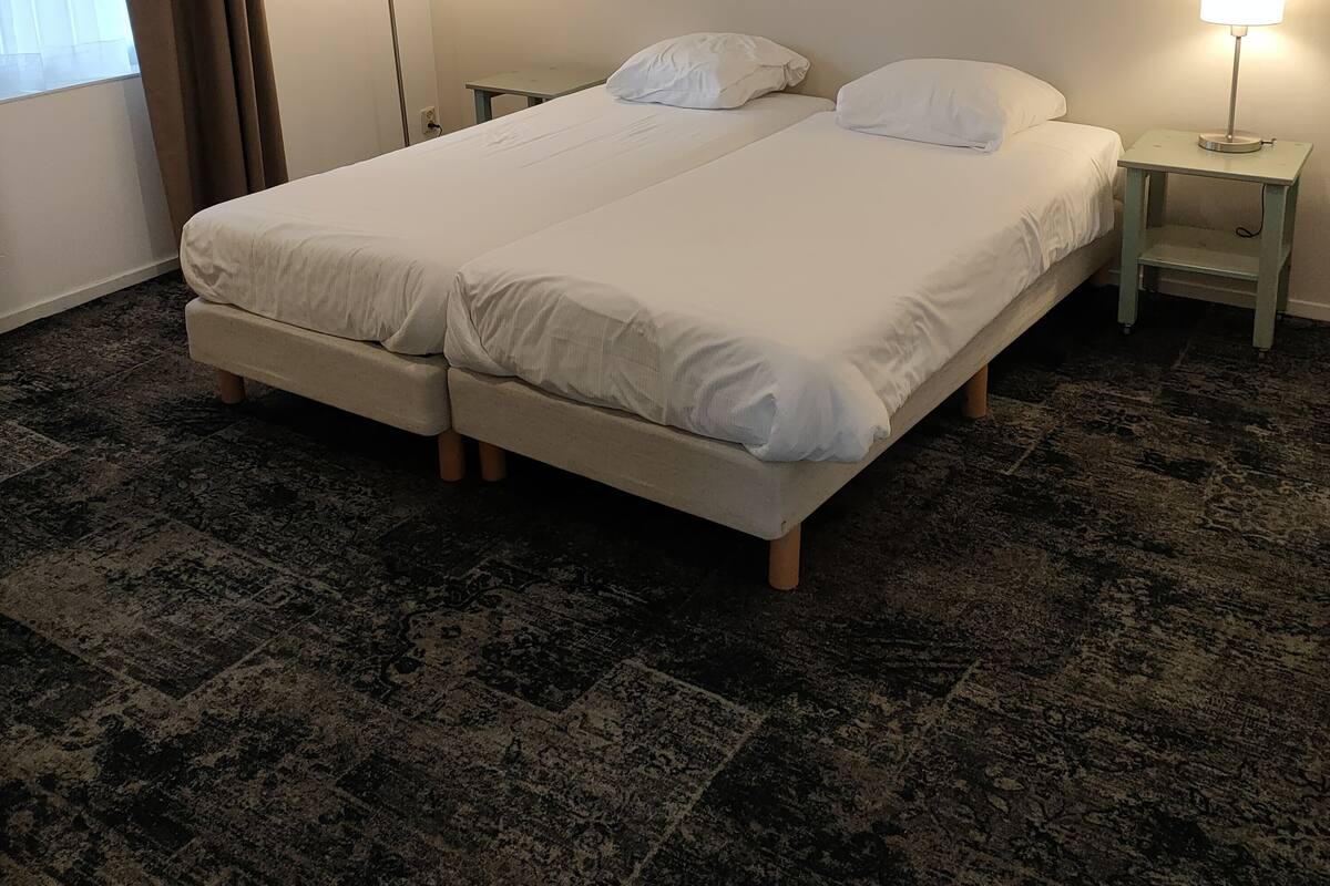 Un lit double avec du linge de lit blanc, deux tables de chevet avec des lampes, un grand tableau floral accroché au mur et une fenêtre avec des rideaux.