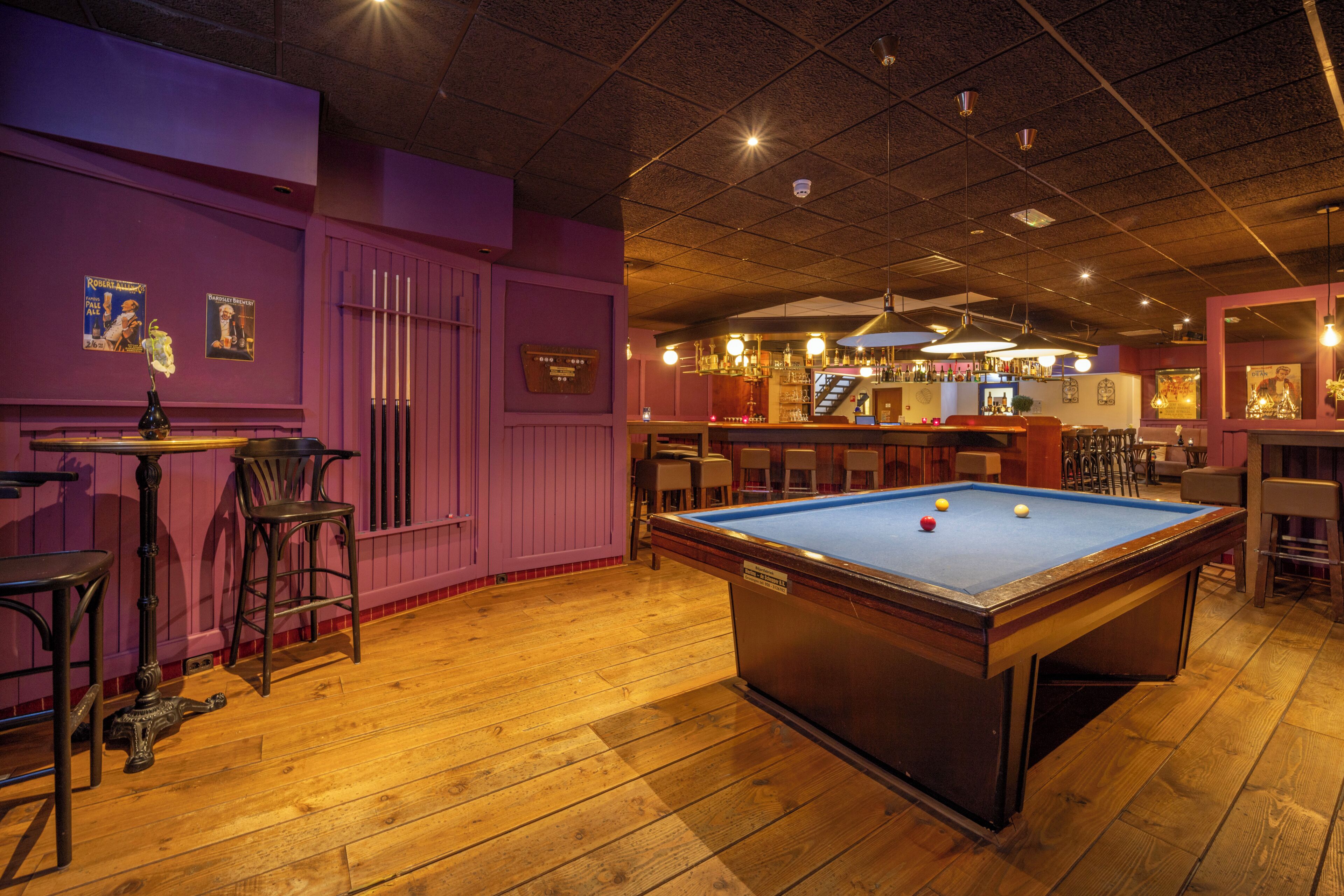 Une table de billard avec un porte-queues et des chaises, un coin bar avec des bouteilles et des verres, et un mur décoré de posters.