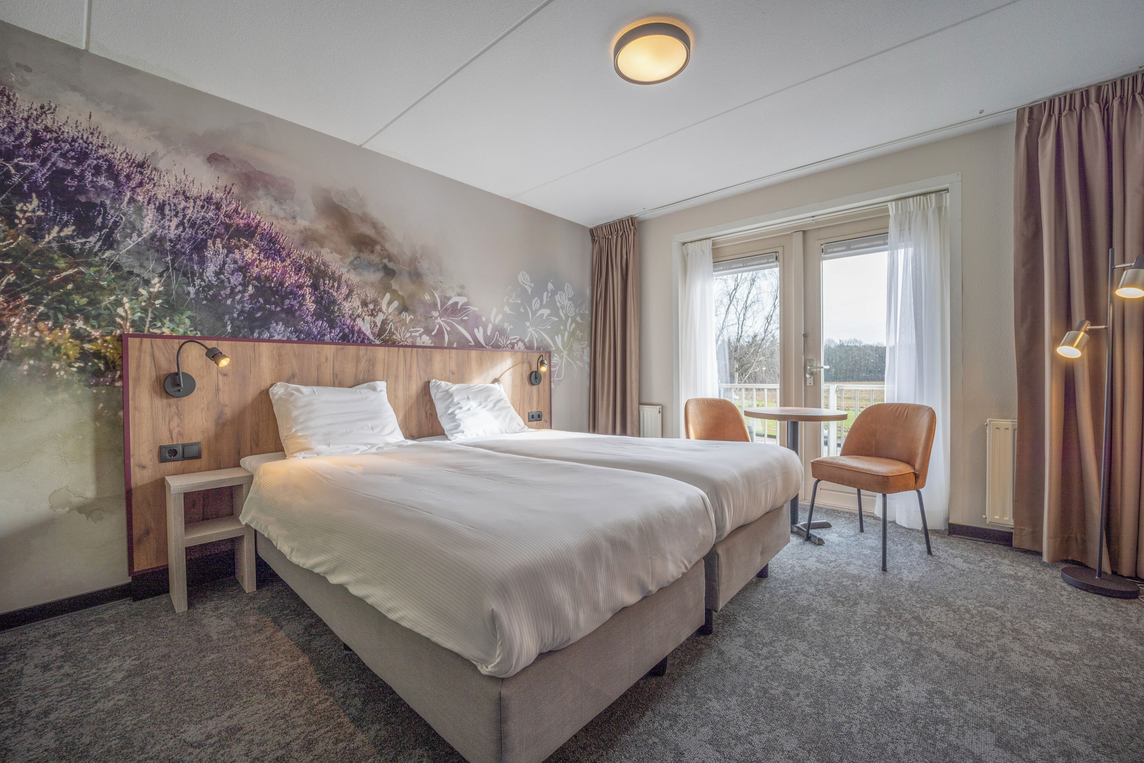 A hotel room with two beds, a small table, a chair, a painting on the wall, and a view through the window.