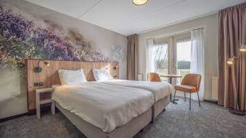 A hotel room with two beds, a small table, a chair, a painting on the wall, and a view through the window.