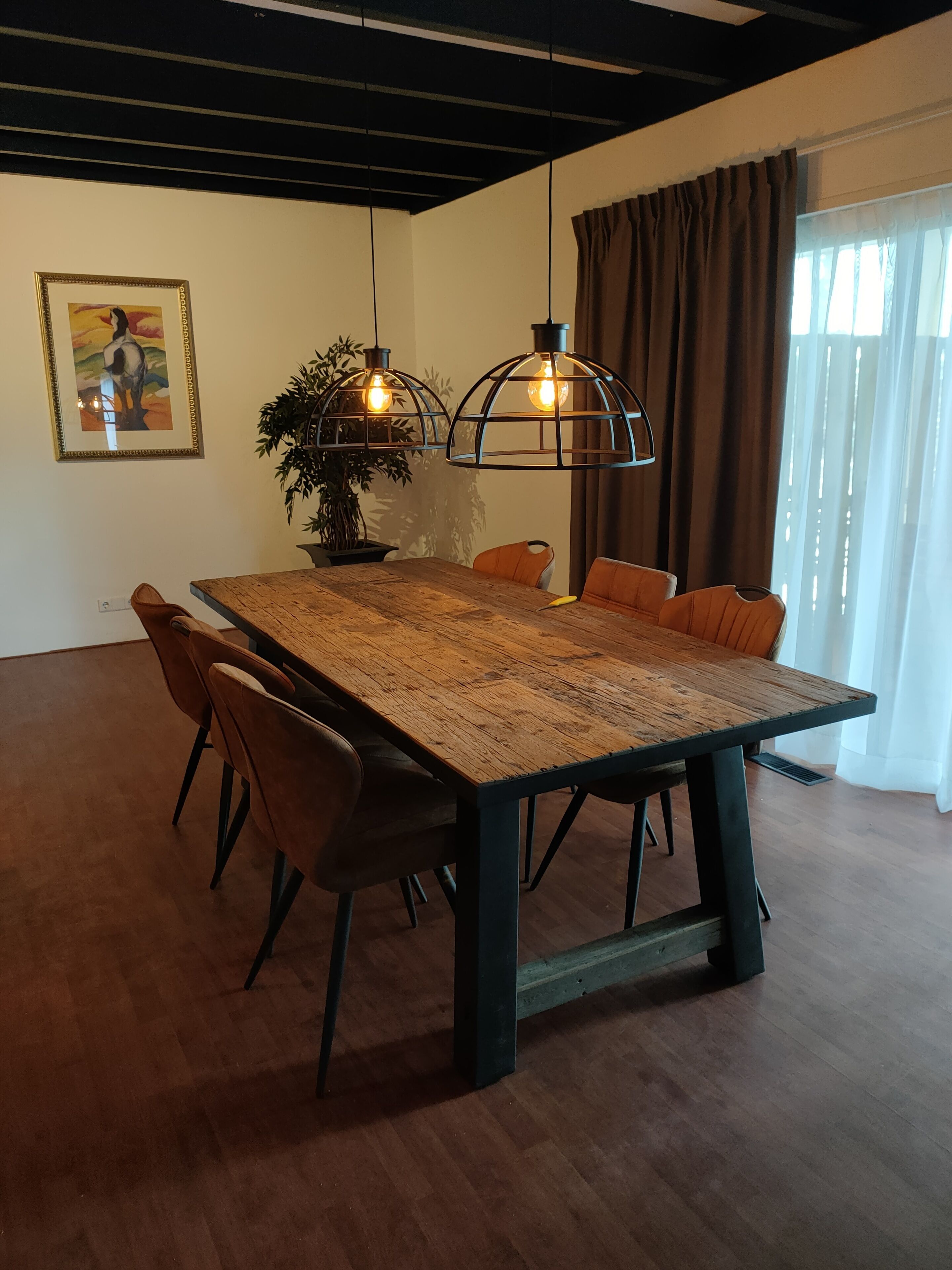 Un comedor con una mesa de madera, sillas, lámparas colgantes, una planta en maceta y un cuadro en la pared.
