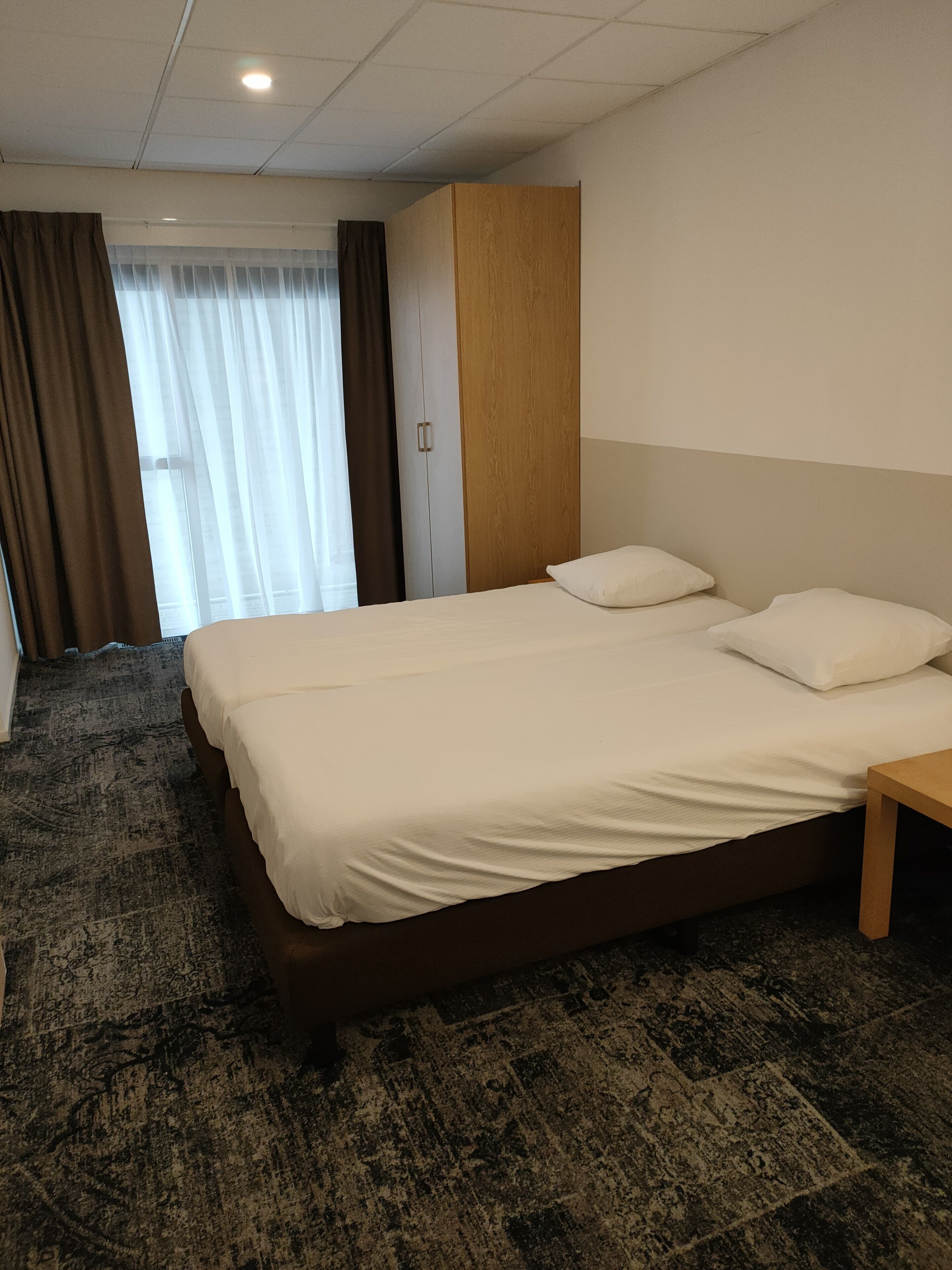 A hotel room with a large bed, a wooden headboard, a small bedside table, and a window with curtains.