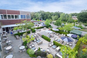 Een terras buiten met verschillende zithoeken, inclusief tafels en stoelen, omringd door groen en parasols.