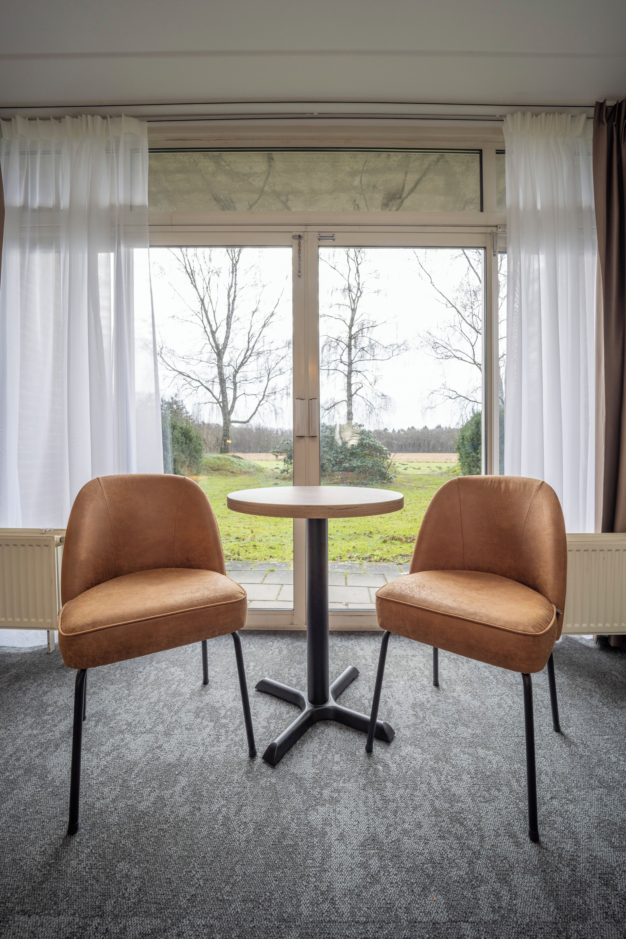 Una habitación con dos sillones de cuero, una mesita redonda y un gran ventanal con cortinas.