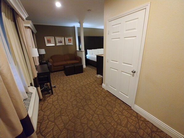 A hotel room with a patterned carpet, a brown sofa, a small table, a white door, and framed pictures on the wall.