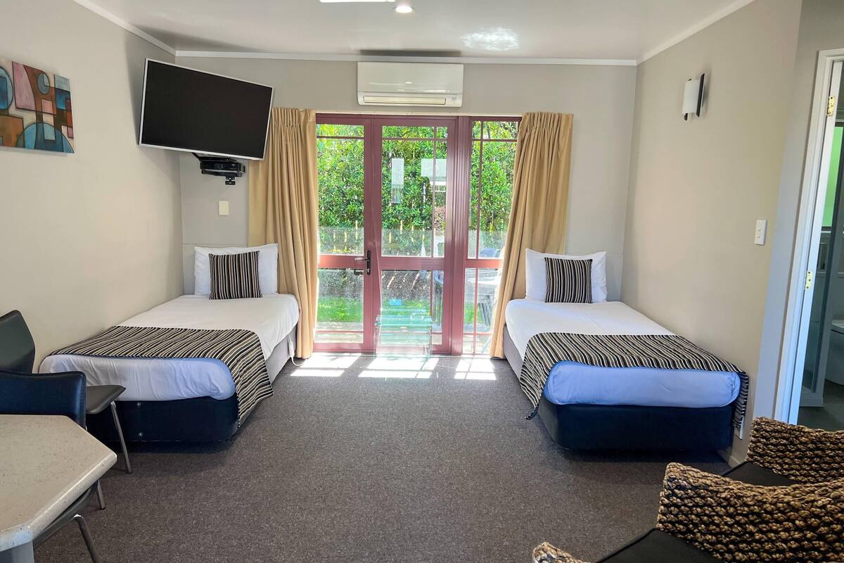 Een hotelkamer met twee bedden, een televisie, een airconditioner en uitzicht op groen door de deur.