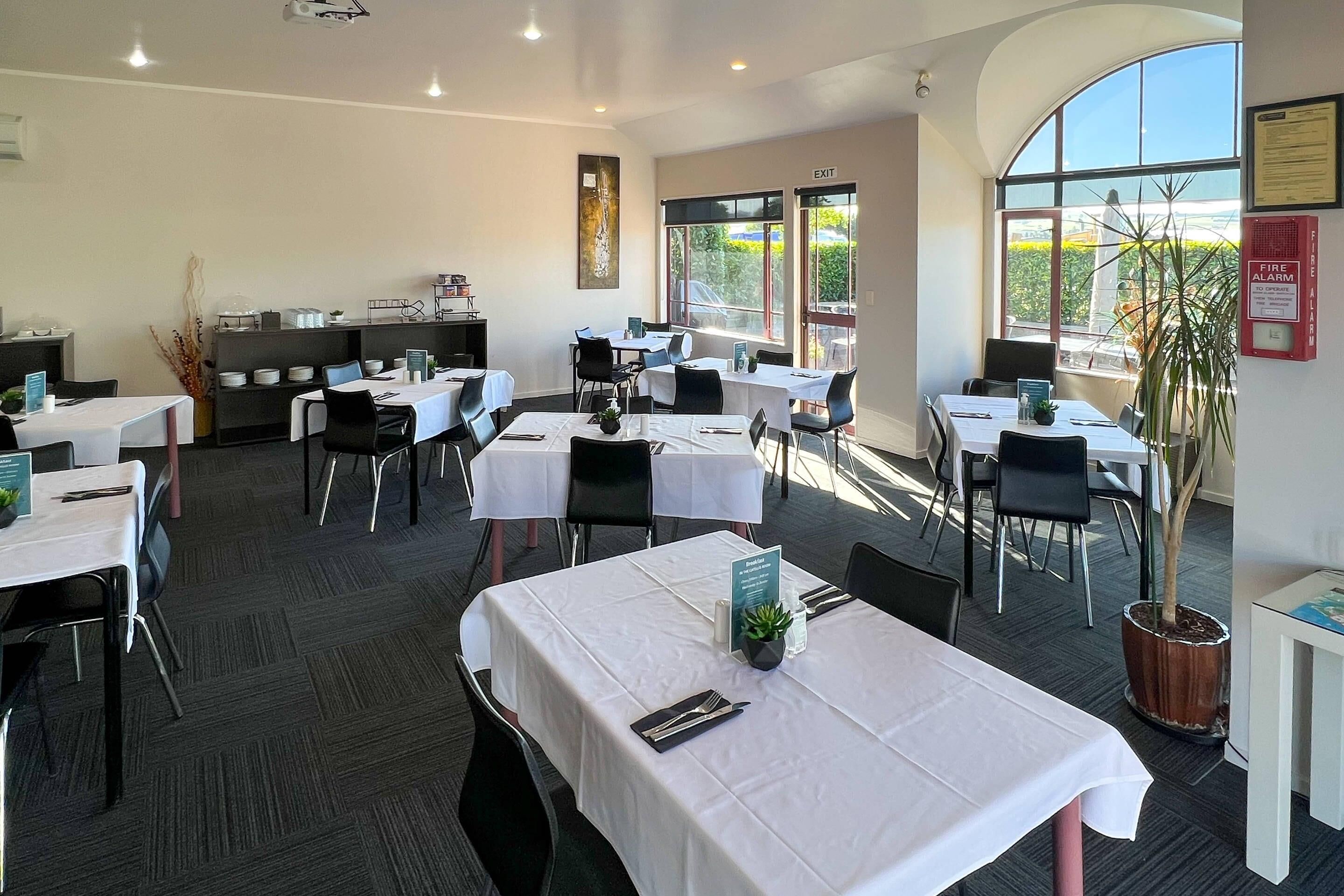 A dining area with tables set for guests, a fire alarm on the wall, and a view of the outdoors through large windows.
