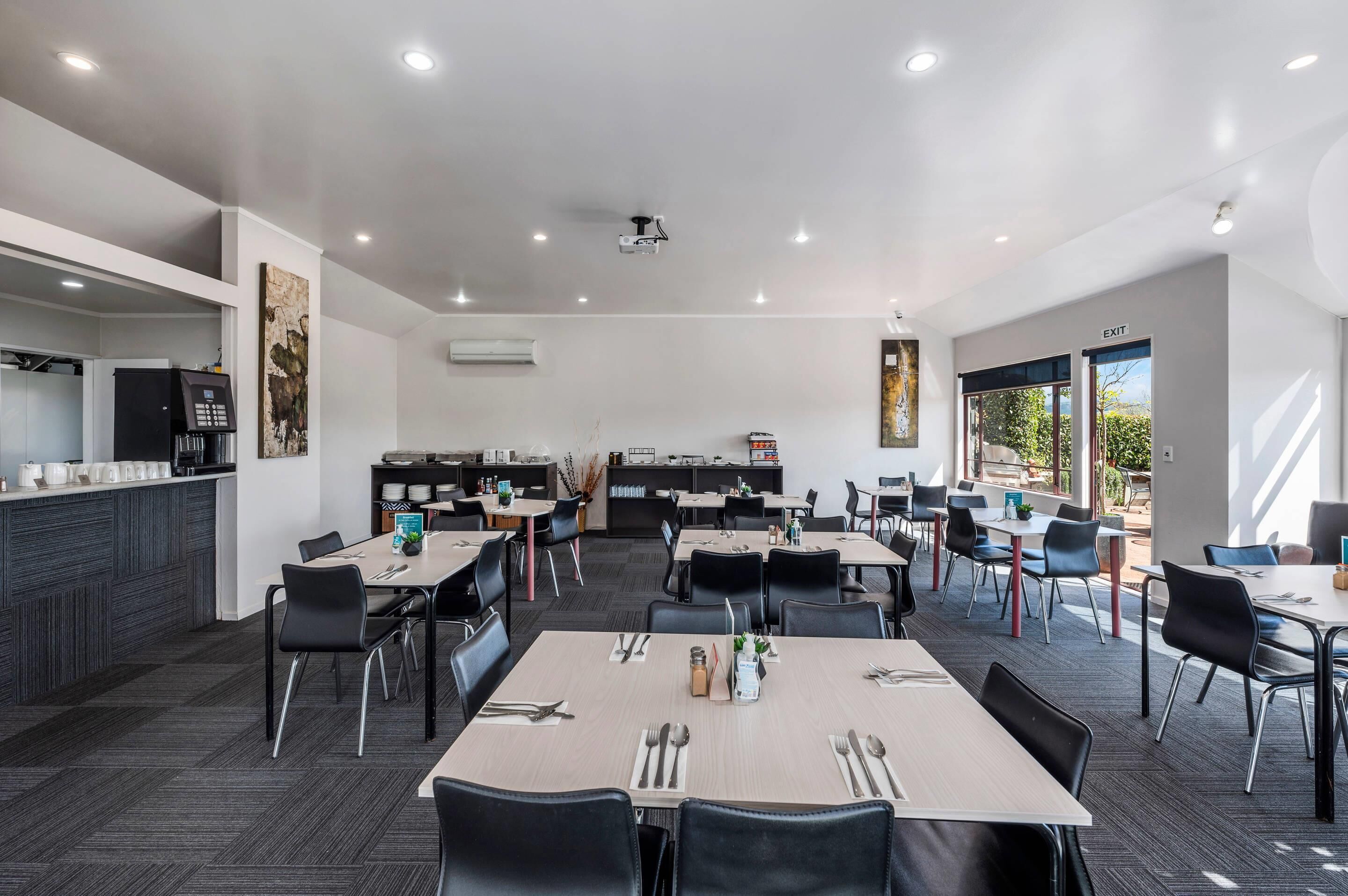 A modern dining area with multiple tables and chairs, a kitchen area, and large windows providing natural light.
