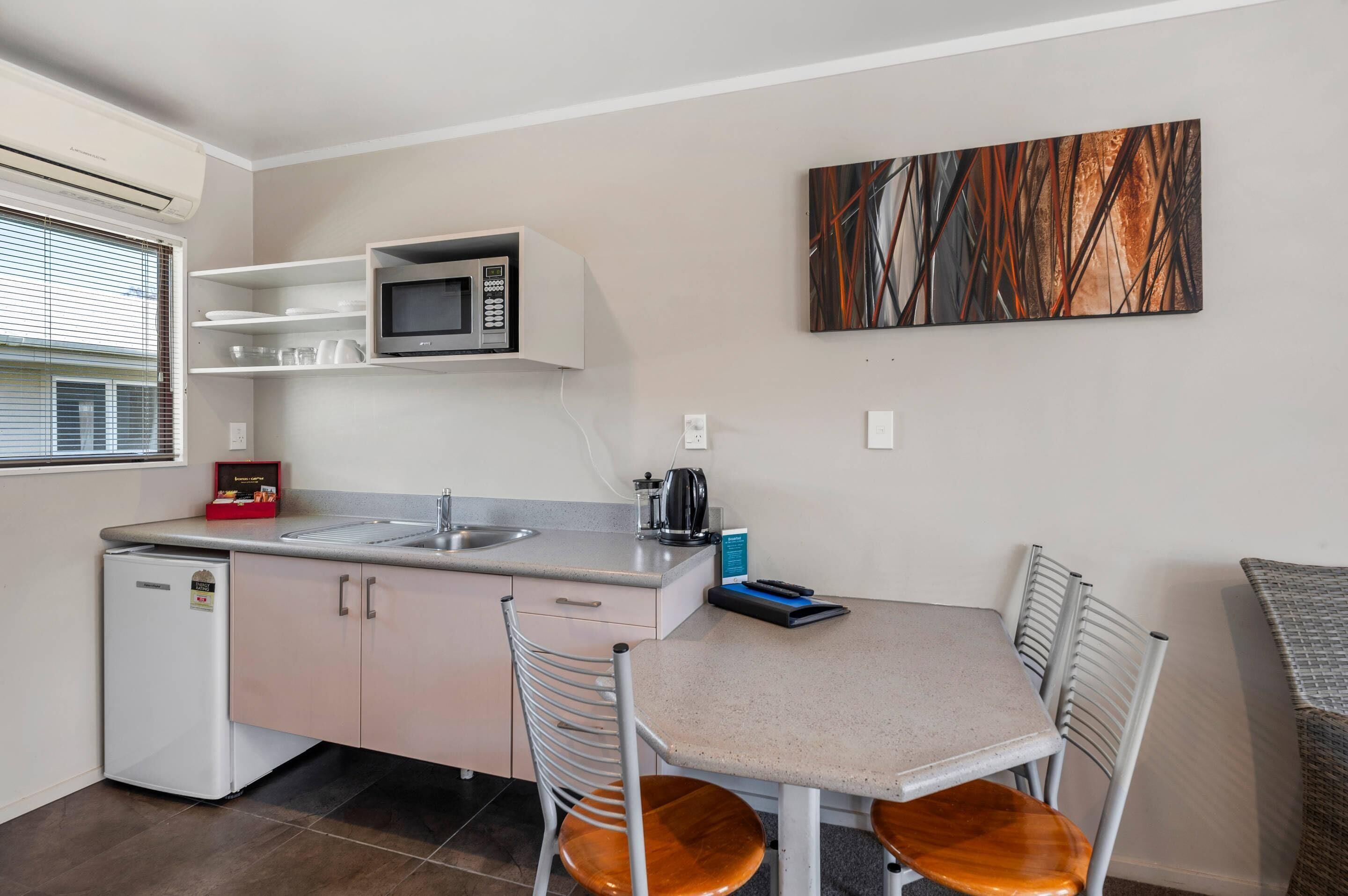 A compact kitchen with a microwave, sink, and a small refrigerator. There is a dining table with chairs and a painting on the wall.