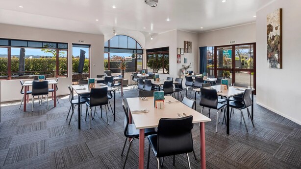A modern conference room with multiple tables and chairs, large windows offering views of the outdoors, and an exit sign on the wall. - Quality Inn Acapulco Taupo (Taupo)