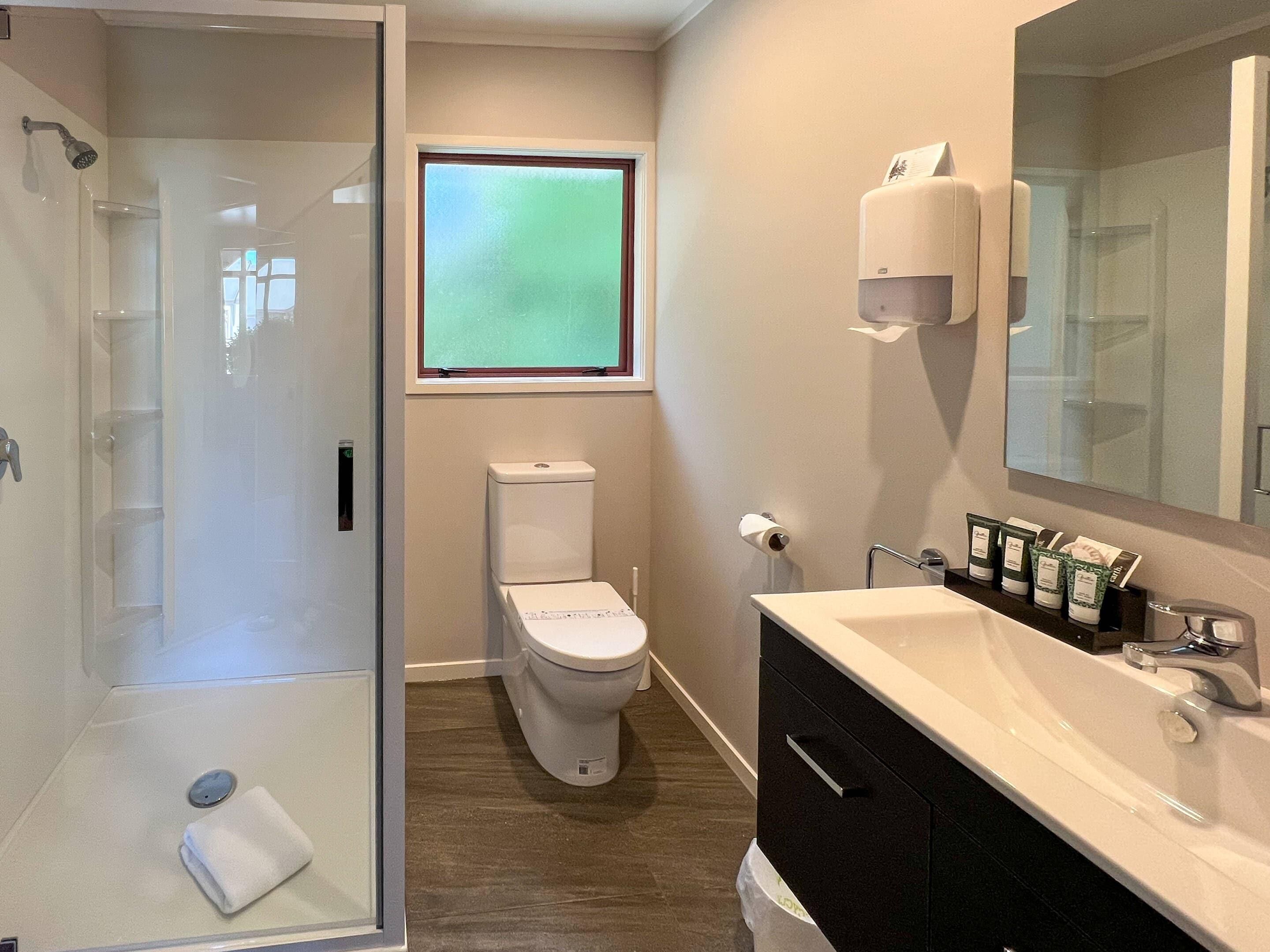 A bathroom with a glass shower enclosure, a toilet, a sink with toiletries, and a window providing natural light.