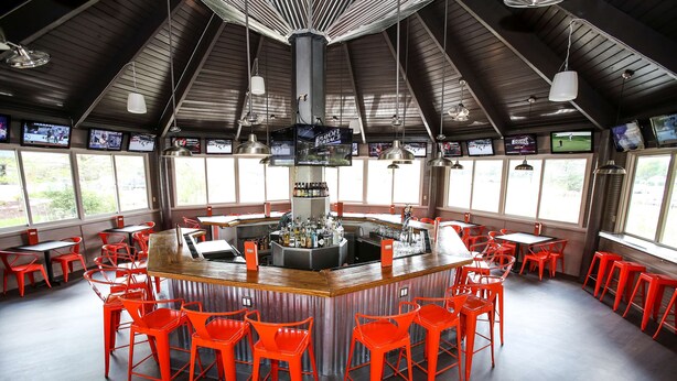 Eine Bar mit einer zentralen Theke, mehreren Bildschirmen und roten Stühlen.