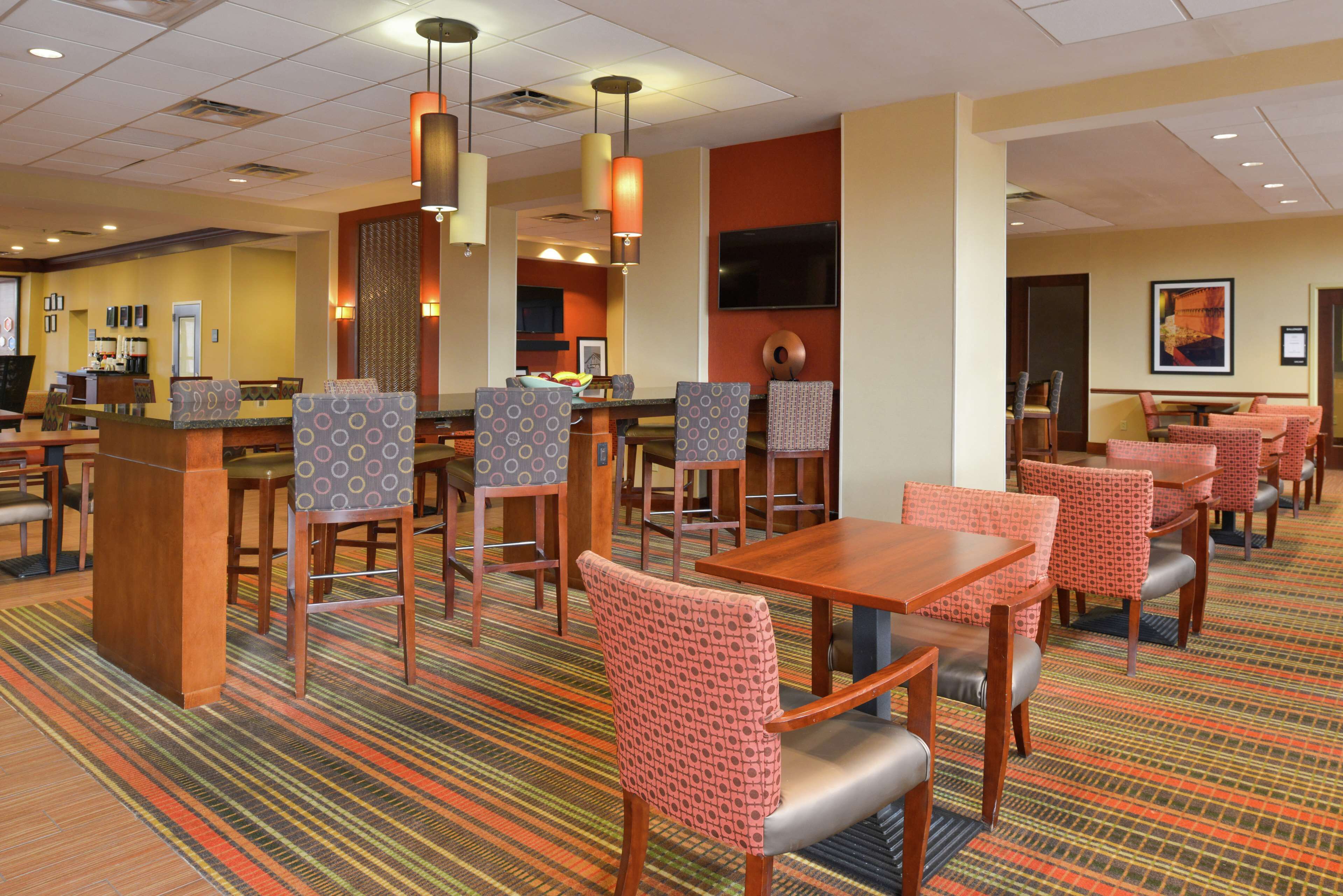A hotel dining area with multiple tables and chairs, a coffee station, and a TV mounted on the wall.