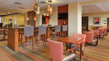 A hotel dining area with multiple tables and chairs, a coffee station, and a TV mounted on the wall.
