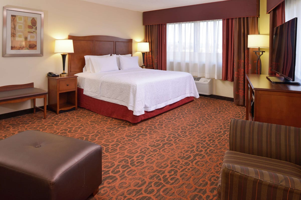 A hotel room with a large bed, two bedside tables with lamps, a chair, a desk with a TV, and a patterned carpet.