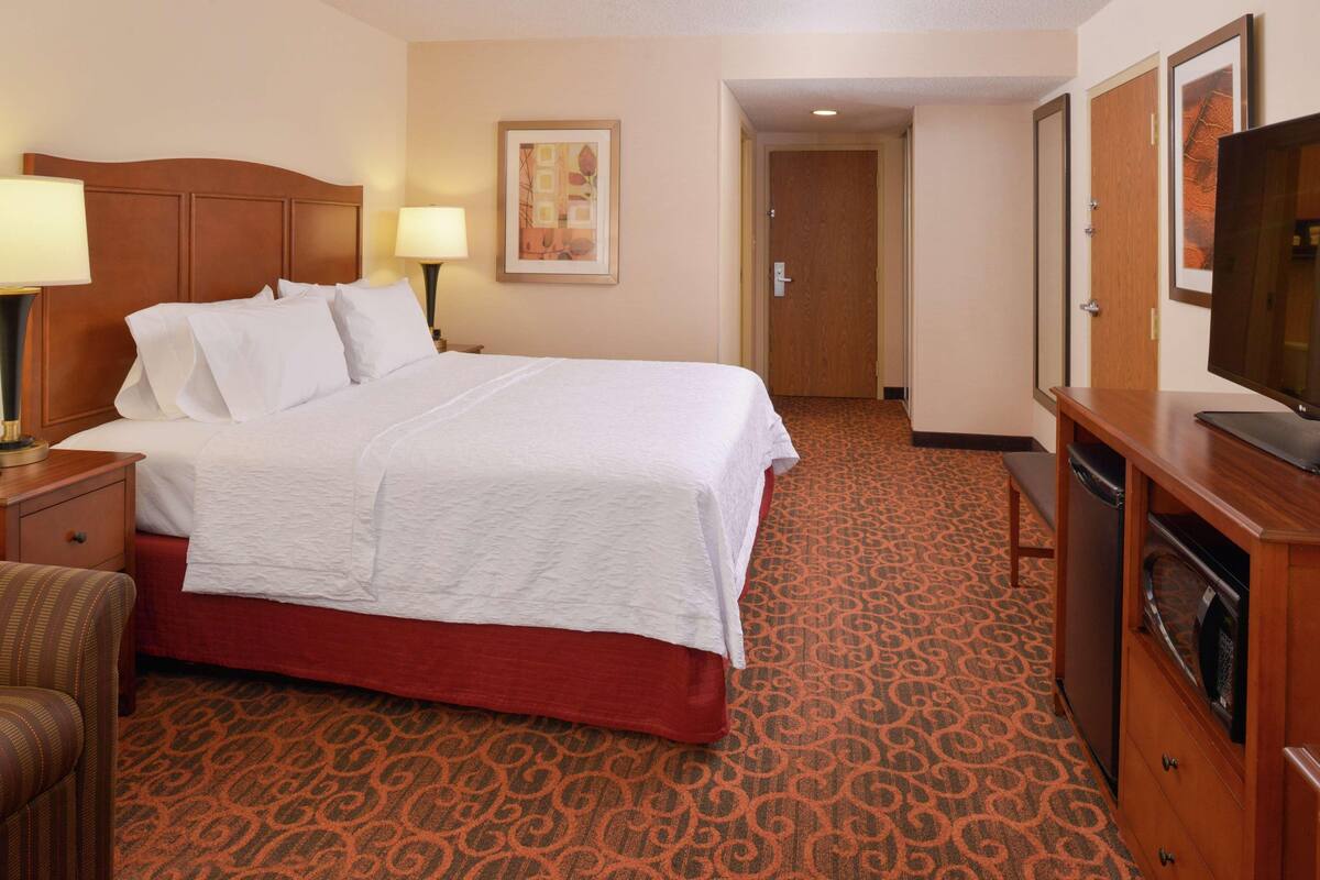 A hotel room with a large bed, a wooden headboard, a desk with a lamp, a television, and a patterned carpet.