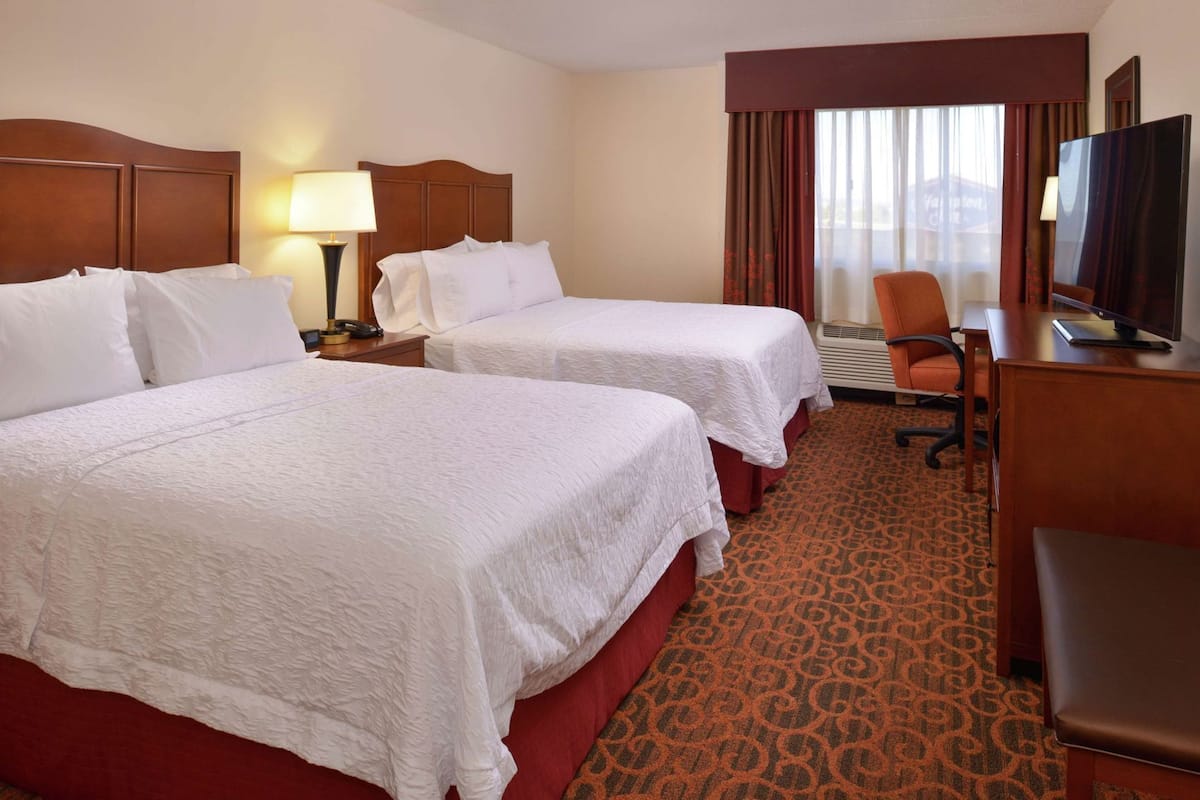 A hotel room with two beds, a desk with a television, a chair, and a window with curtains.
