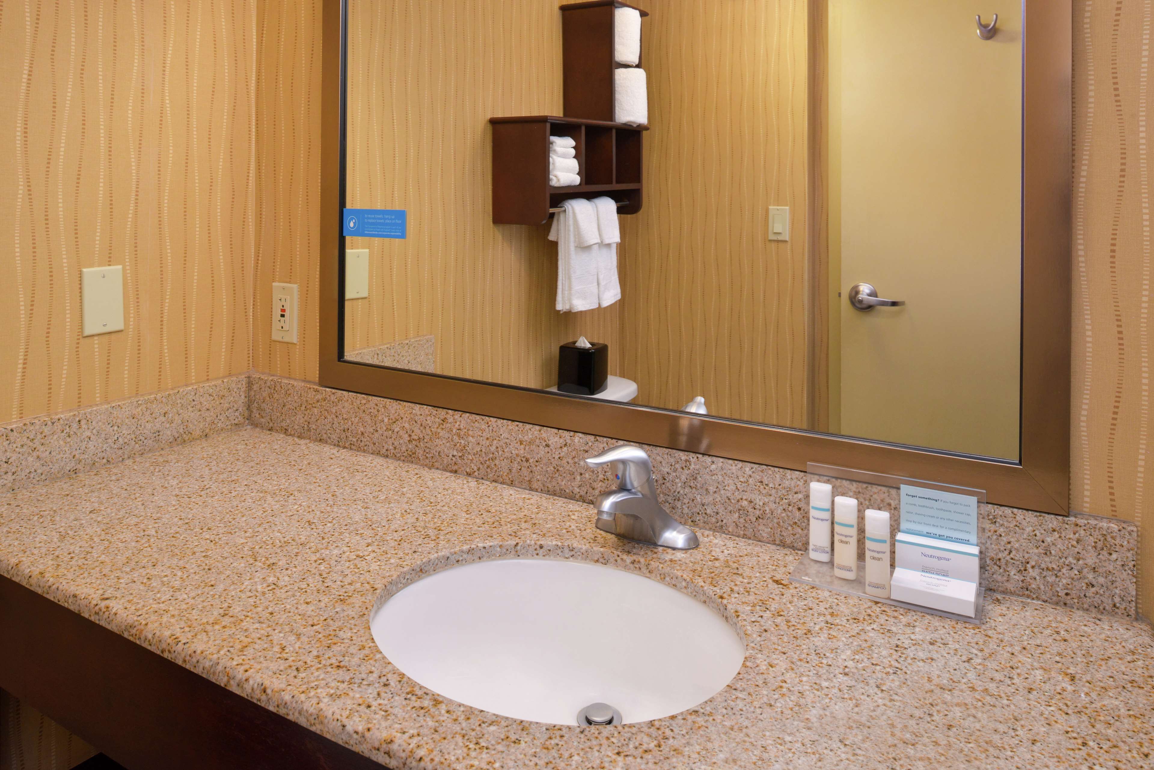 A hotel bathroom with a granite countertop, a white sink, a mirror, a towel rack, and a soap dispenser.