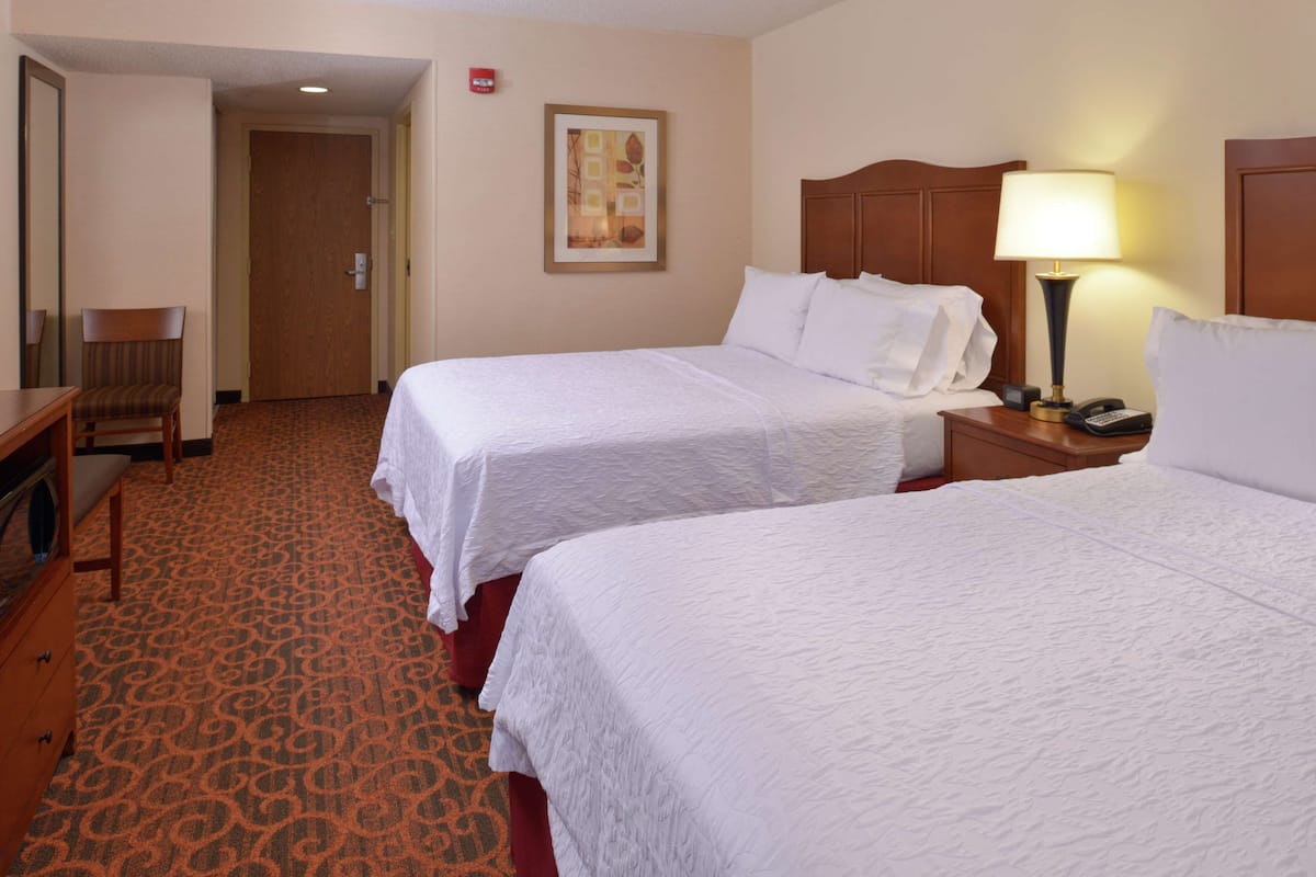 A hotel room with two single beds, a desk, a chair, and a nightstand with a lamp.
