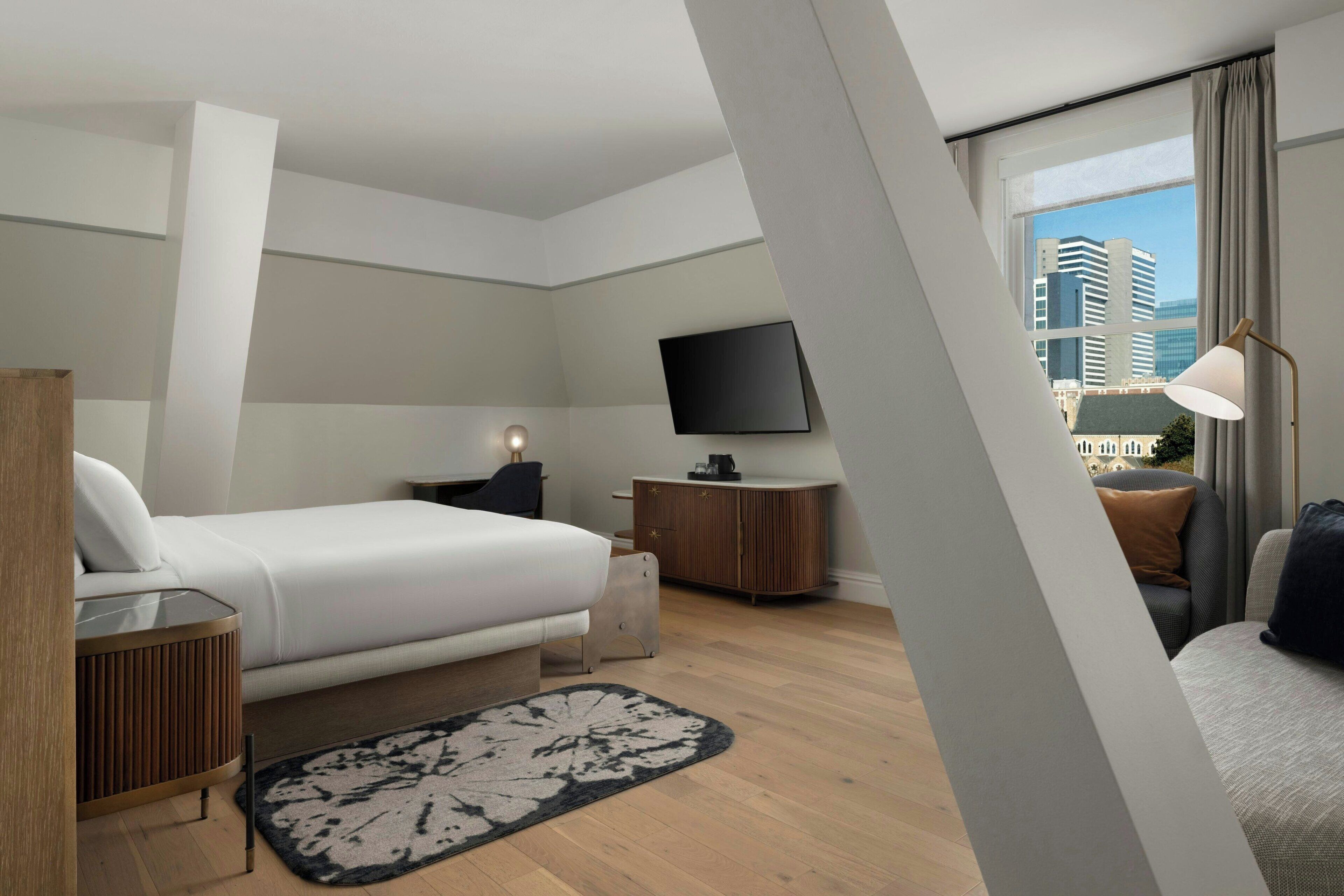 Una habitación de hotel moderna con una cama grande, un televisor de pantalla plana en la pared, una mesita de madera y vistas a la ciudad a través de la ventana.