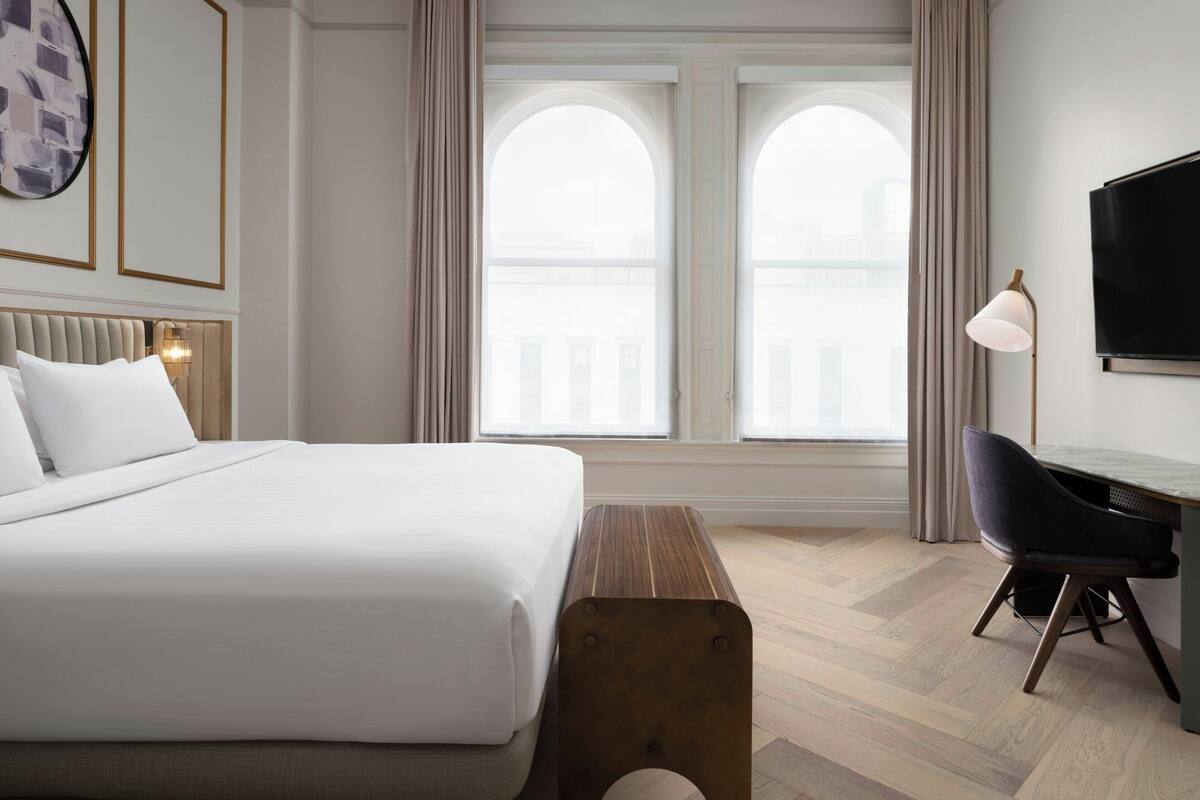 Ein Hotelzimmer mit einem großen Bett, einem Schreibtisch mit Stuhl, einem Fernseher und einem Fenster mit Vorhängen.
