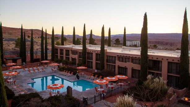 A hotel pool area with sun loungers and umbrellas, surrounded by tall cypress trees and a view of distant mountains.