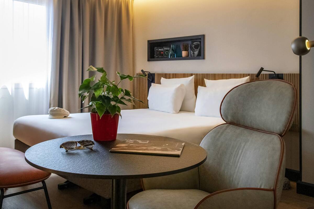 A hotel room with a bed, a bedside table with a plant, a chair, and a lamp.