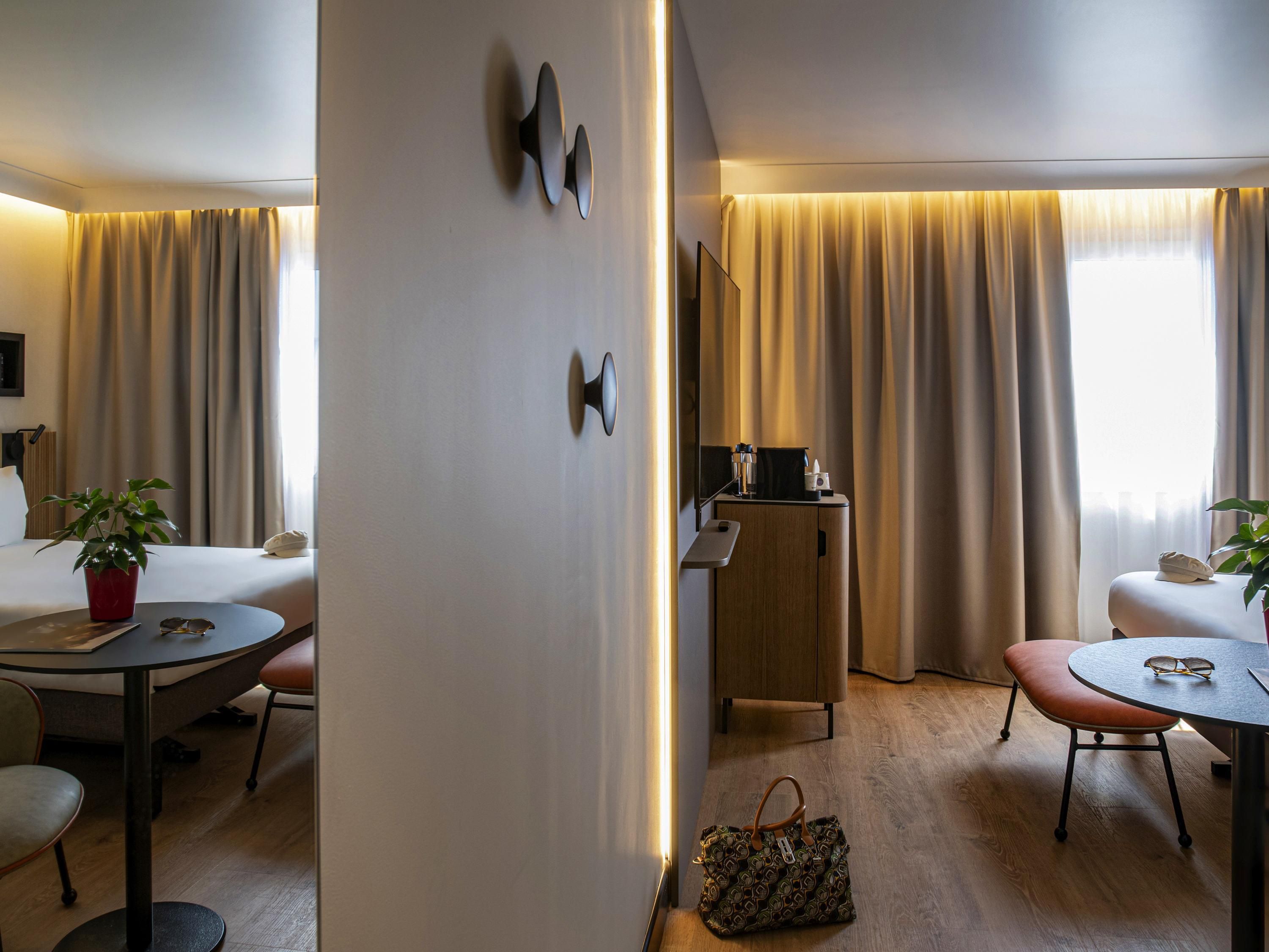 Hotellihuoneessa on sänky, työpöytä, tuoli, pieni pöytä, kasvi ja lattialla käsilaukku.