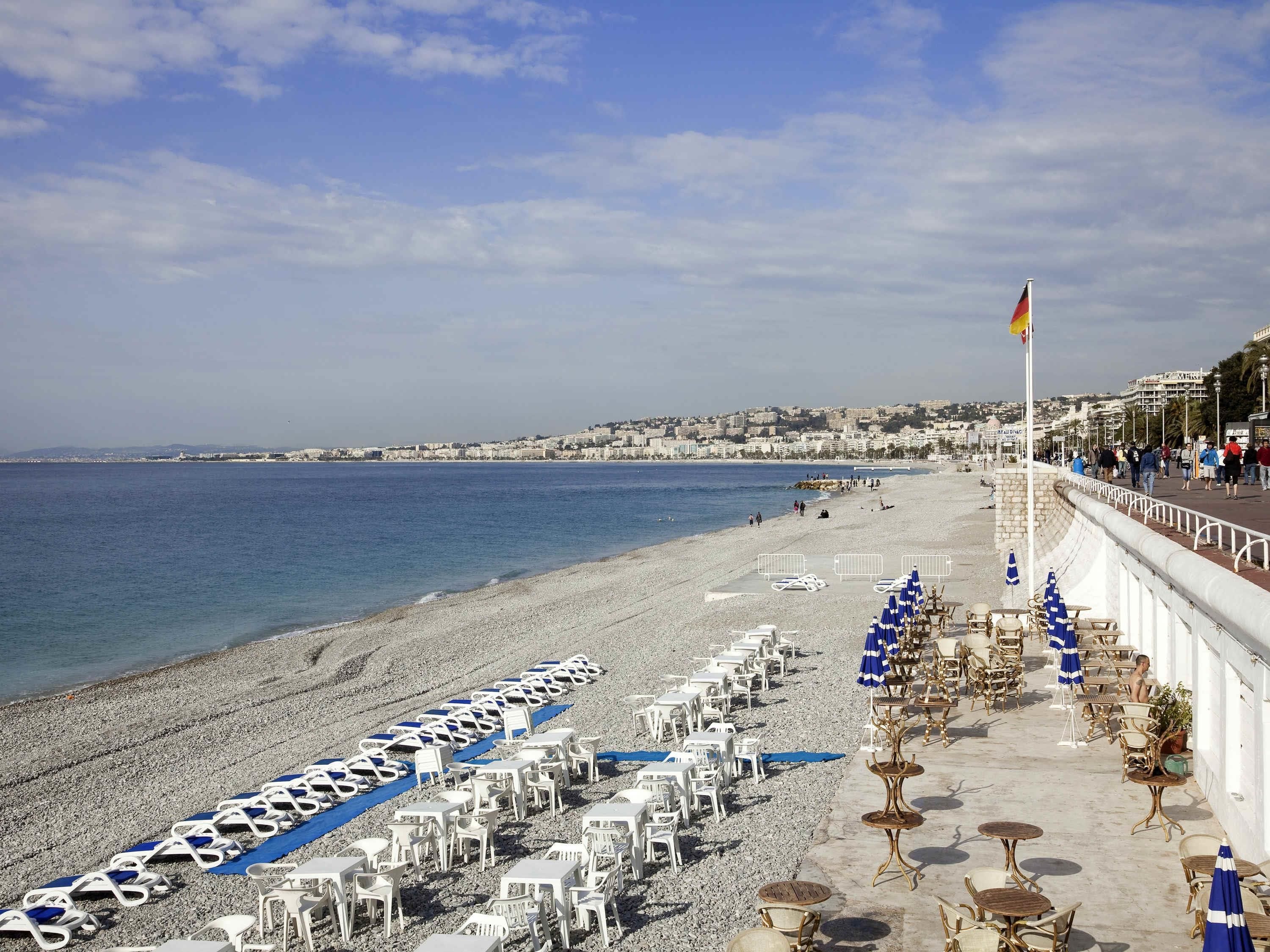 Un hotel frente a la playa con filas de tumbonas y mesas, una bandera y vistas al mar y al paisaje urbano.
