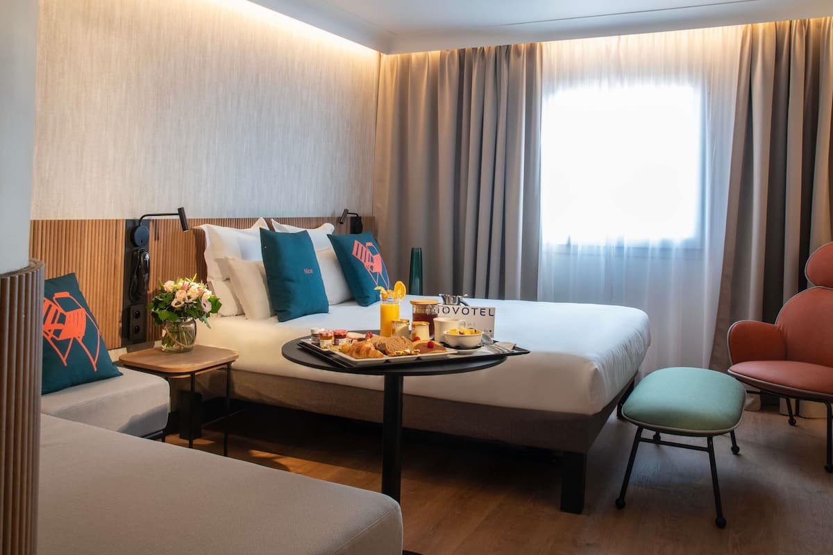 A hotel room with a bed, a small table with breakfast, a chair, and a desk with a phone.