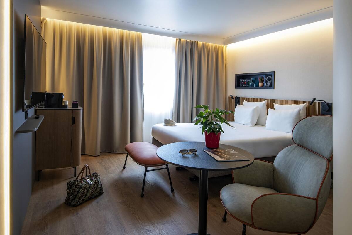 A hotel room with a bed, a desk, a chair, a table, a TV, and a window with curtains.