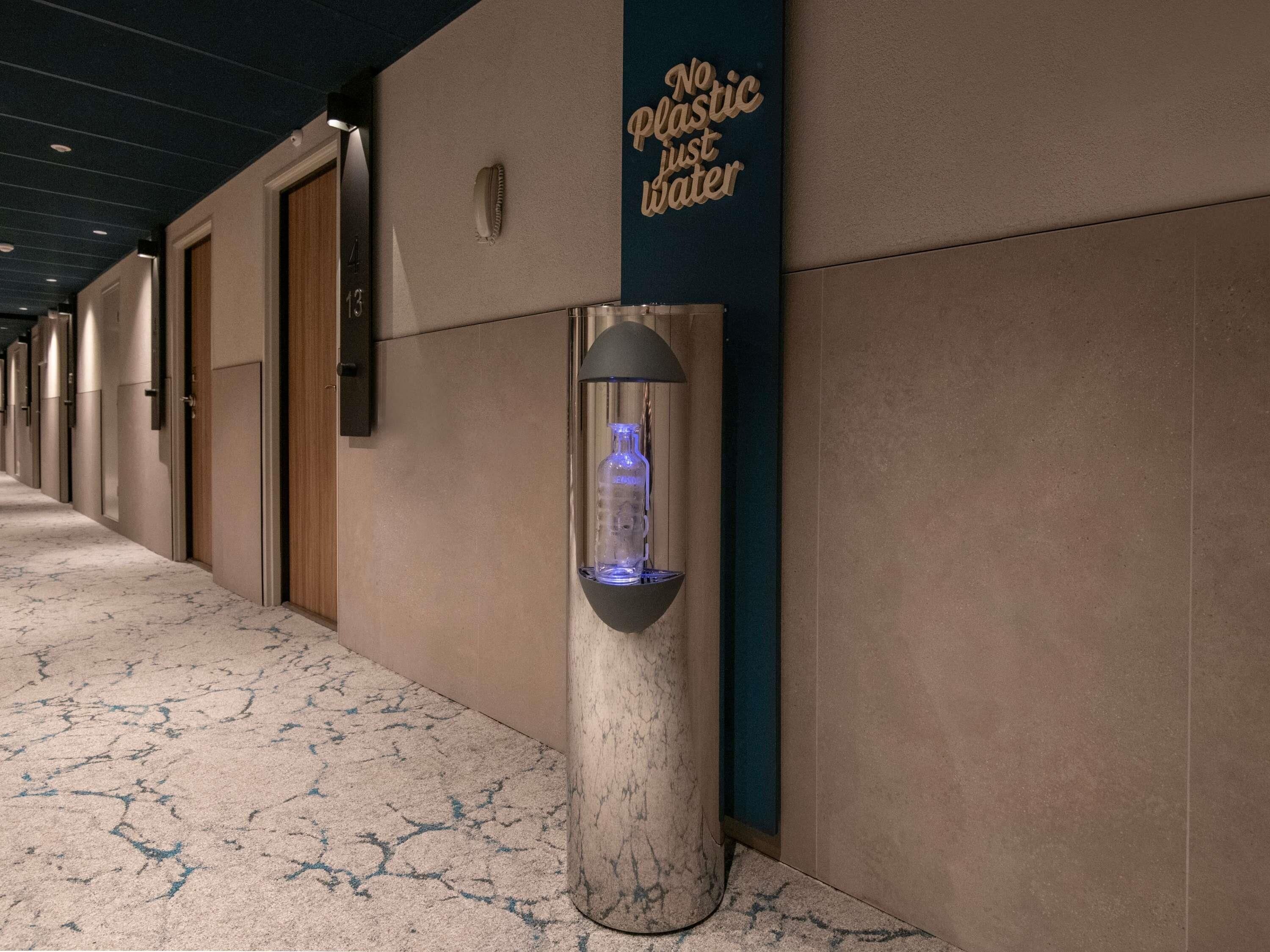 Un pasillo con un dispensador de agua fijado a un pilar, un cartel en la pared que dice “No Plastic Just Water”, y puertas a la izquierda.