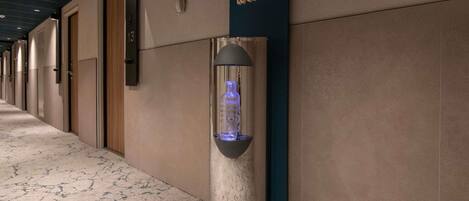 Un pasillo con un dispensador de agua fijado a un pilar, un cartel en la pared que dice “No Plastic Just Water”, y puertas a la izquierda.
