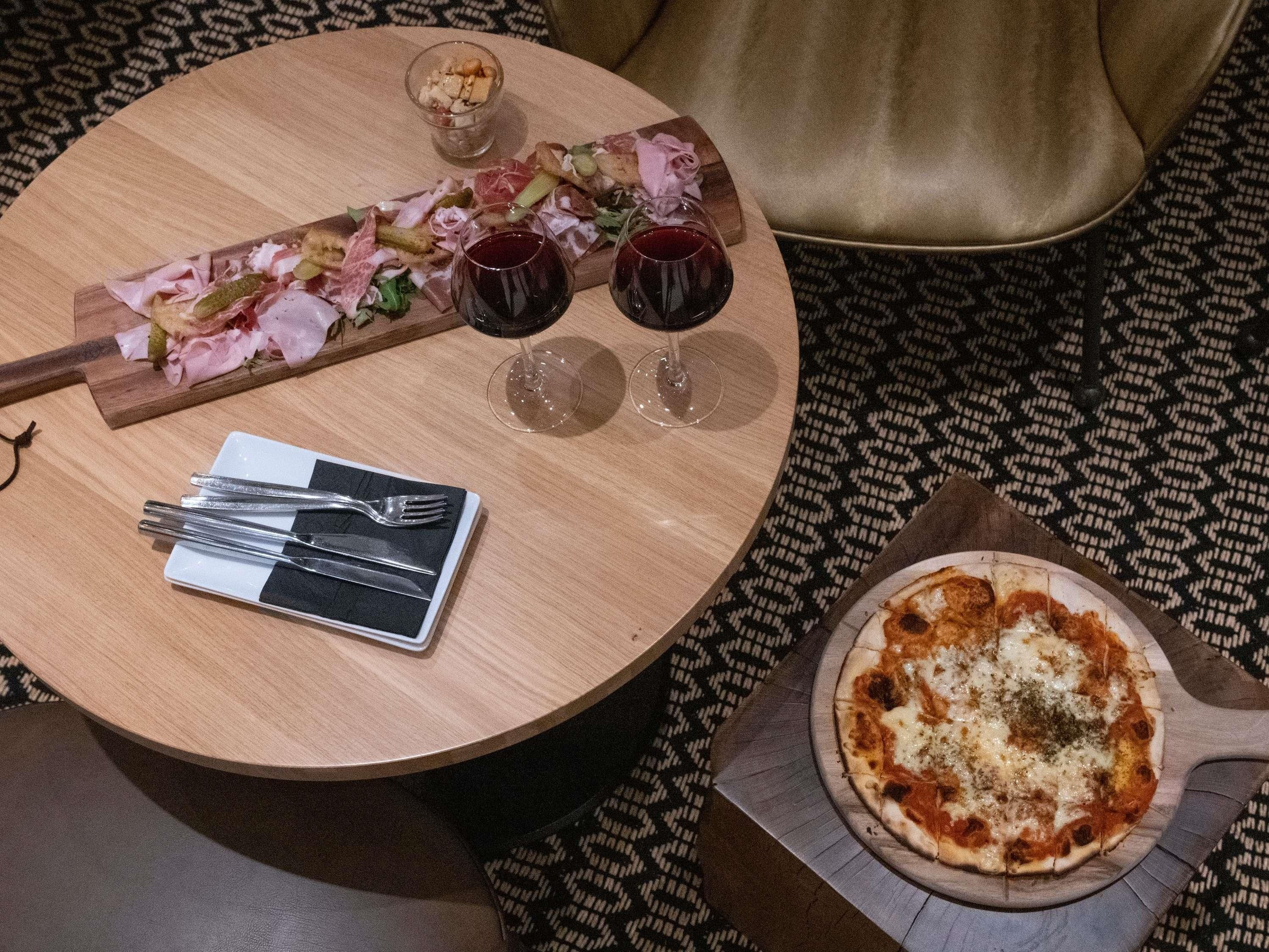 A round wooden table with a pizza, two glasses of red wine, a small bowl of nuts, and a plate of sliced meats and cheeses.
