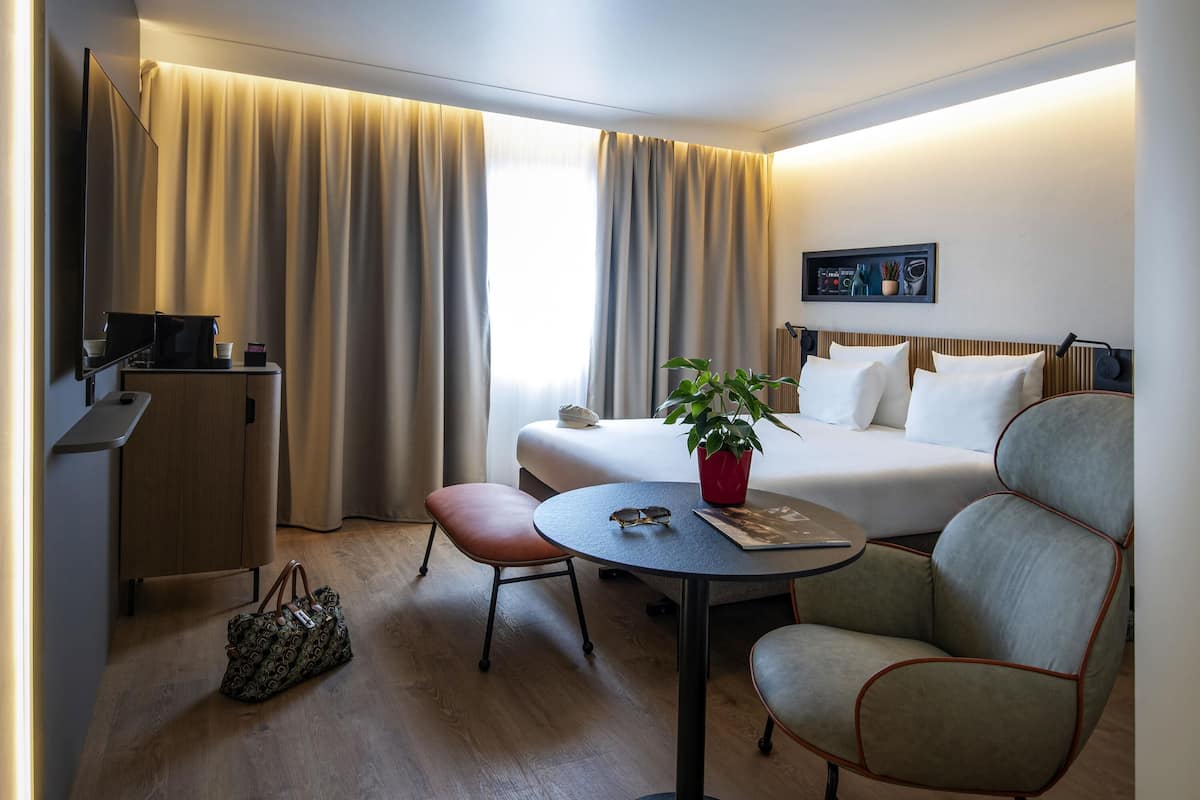 A hotel room with a bed, a desk, a chair, a table, a TV, and a window with curtains.