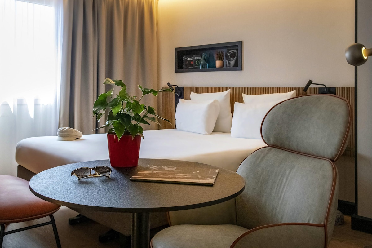Une chambre d’hôtel avec un lit, une table de chevet ornée d’une plante, une chaise et une lampe.