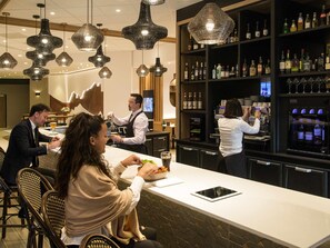 A bar area with a counter, shelves of liquor bottles, and hanging pendant lights. - Novotel Nice Arenas Aeroport (Nice)