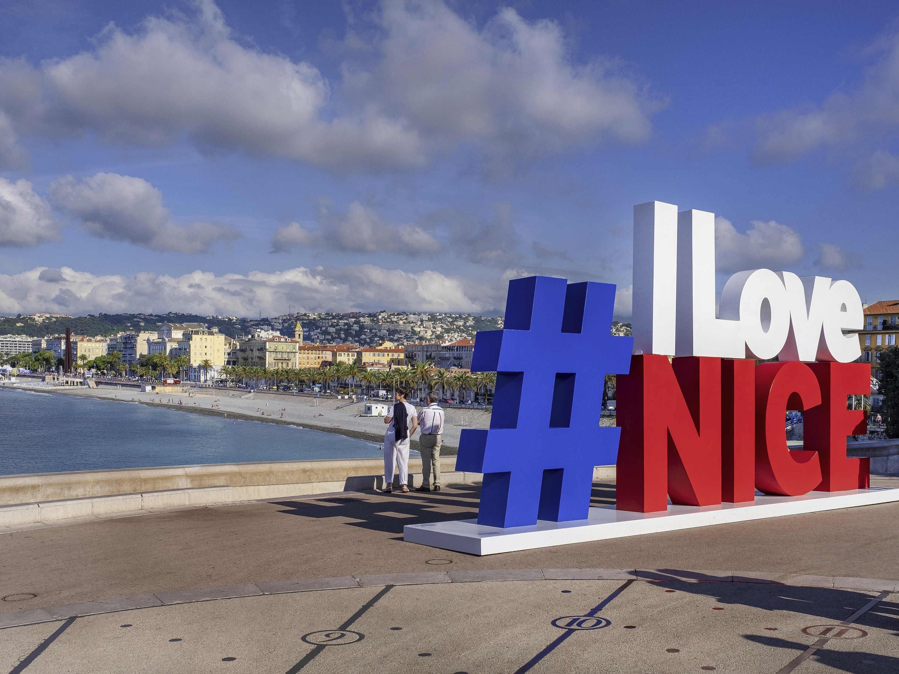 Une vue urbaine côtière avec une grande pancarte bien visible sur laquelle on peut lire « #J’aimeNice », une plage et, au loin, les bâtiments de la ville.