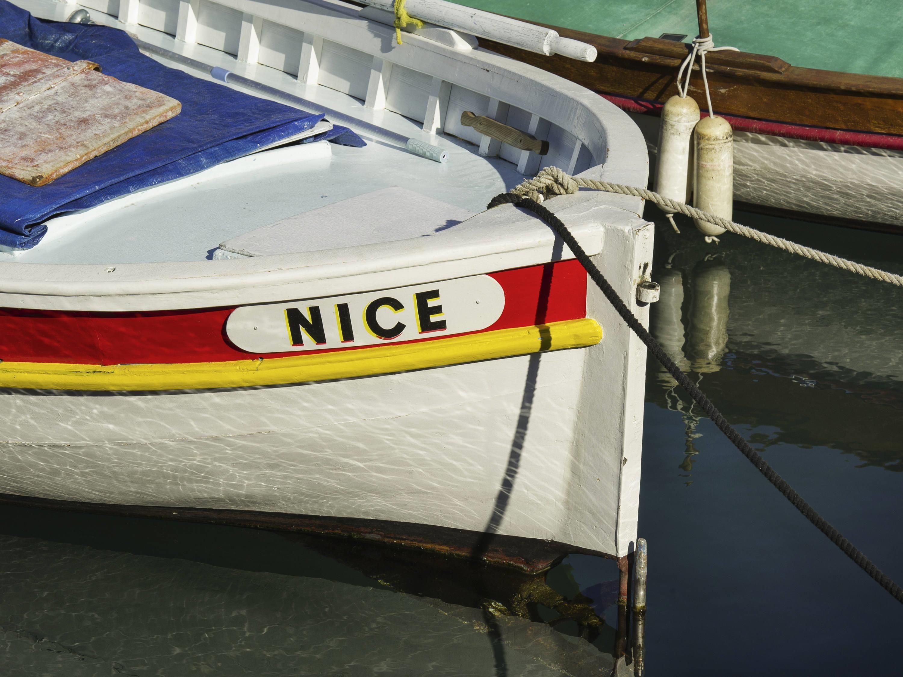 En liten vit båt med namnet ”NICE” målat på sidan, förtöjd vid en brygga med ett rep.