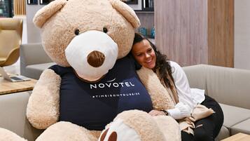 Una persona sentada en un sofá con un gran osito de peluche. El peluche tiene una etiqueta que dice “NOVOTEL” y un hashtag “#TIMEISONYOURSIDE”.