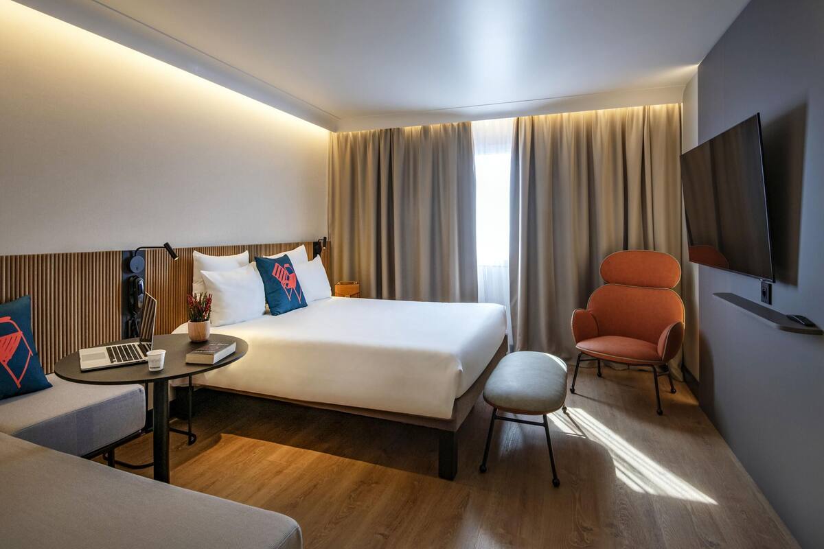 A hotel room with a bed, a desk with a laptop, a chair, a TV, and a window with curtains.