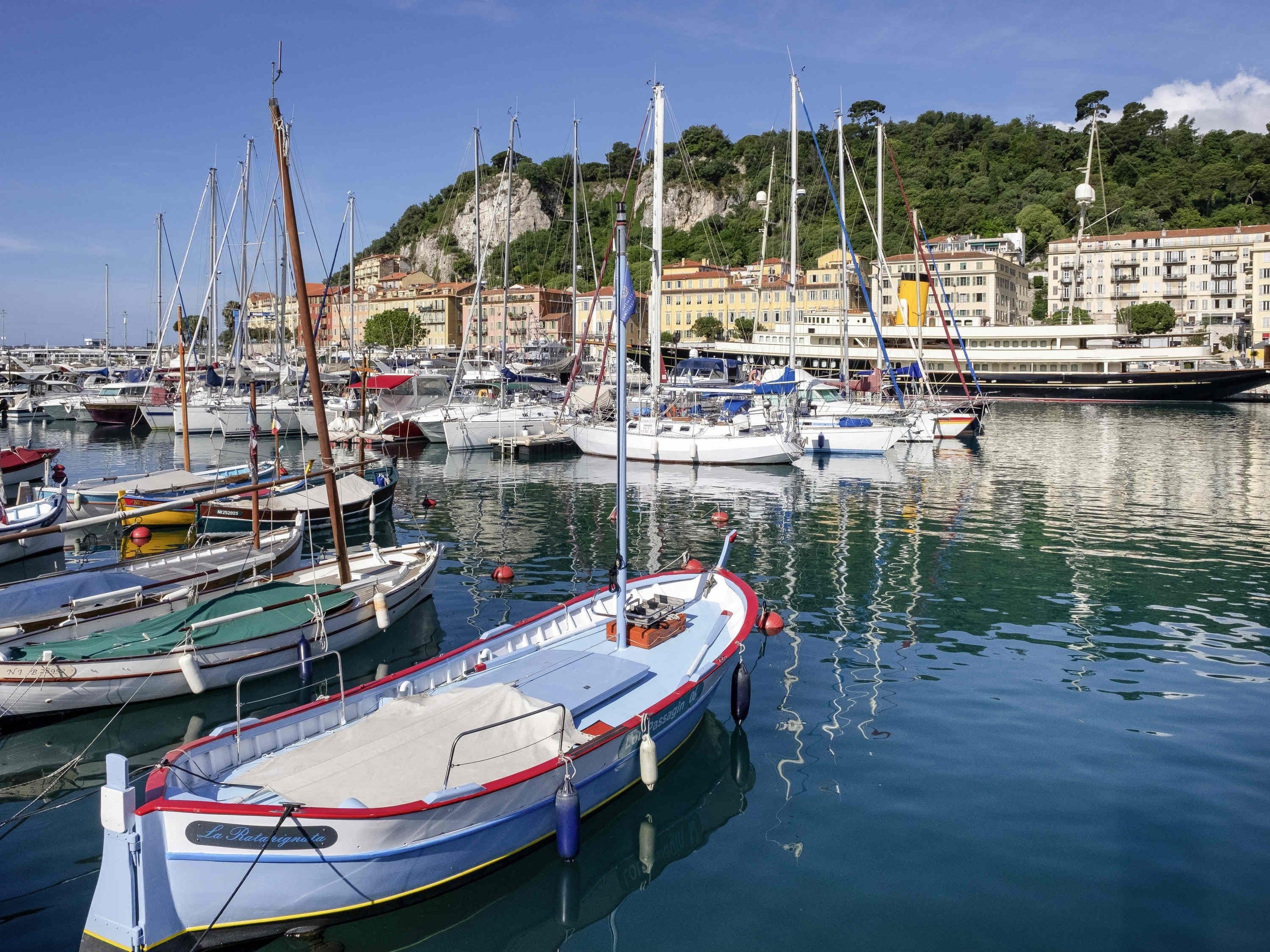 A marina with various boats docked, a hill with buildings in the background, and calm water reflecting the boats.