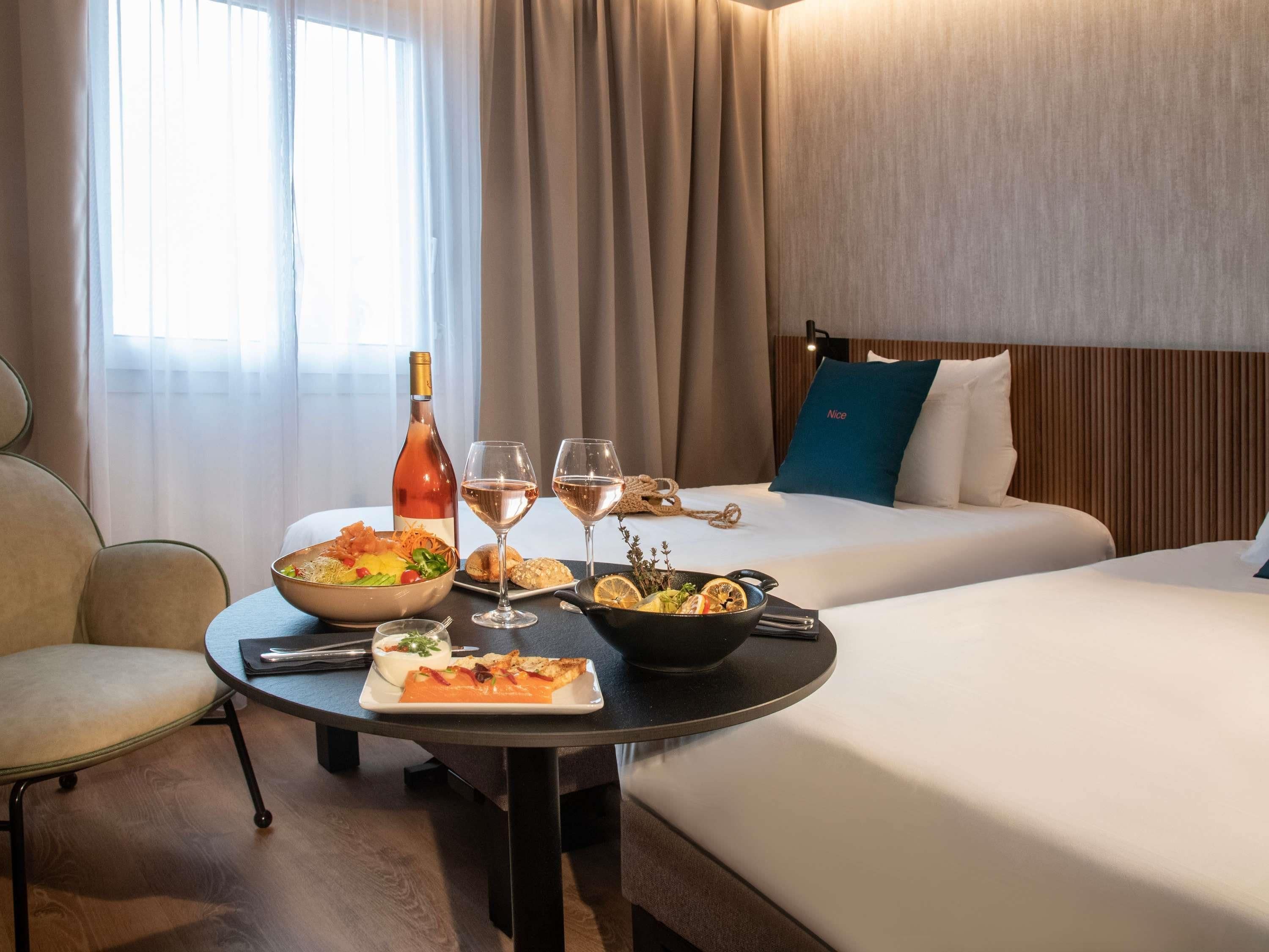 A hotel room with a dining table set with food and drinks, a bed with a blue pillow, and a window with curtains.